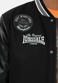 Svart varsityjacka med läderärmar. Har broderade lappar, inklusive "Heavy Weight Kings" och "The Original Lonsdale" logotyper.