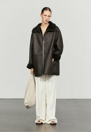 Frau trägt eine übergroße dunkelbraune Jacke mit Reißverschluss, Pelzkragen und -manschetten, weiße weite Hosen, hält eine beige Tasche und steht auf einem grauen Boden.
