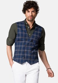 Granatowa kamizelka w kratę z dekoltem w kształcie litery V, podwójnymi guzikami i dwiema przednimi kieszeniami, w zestawieniu z ciemnozieloną koszulą i białymi spodniami.