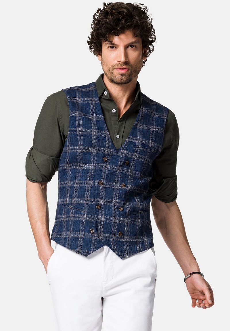 Granatowa kamizelka w kratę z dekoltem w kształcie litery V, podwójnymi guzikami i dwiema przednimi kieszeniami, w zestawieniu z ciemnozieloną koszulą i białymi spodniami.