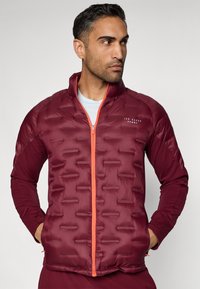 Hnedý vypchávaný kabát s textúrovaným povrchom, oranžovým zipsom a priliehavým dizajnom. Má stojací golier a logo na hrudi.