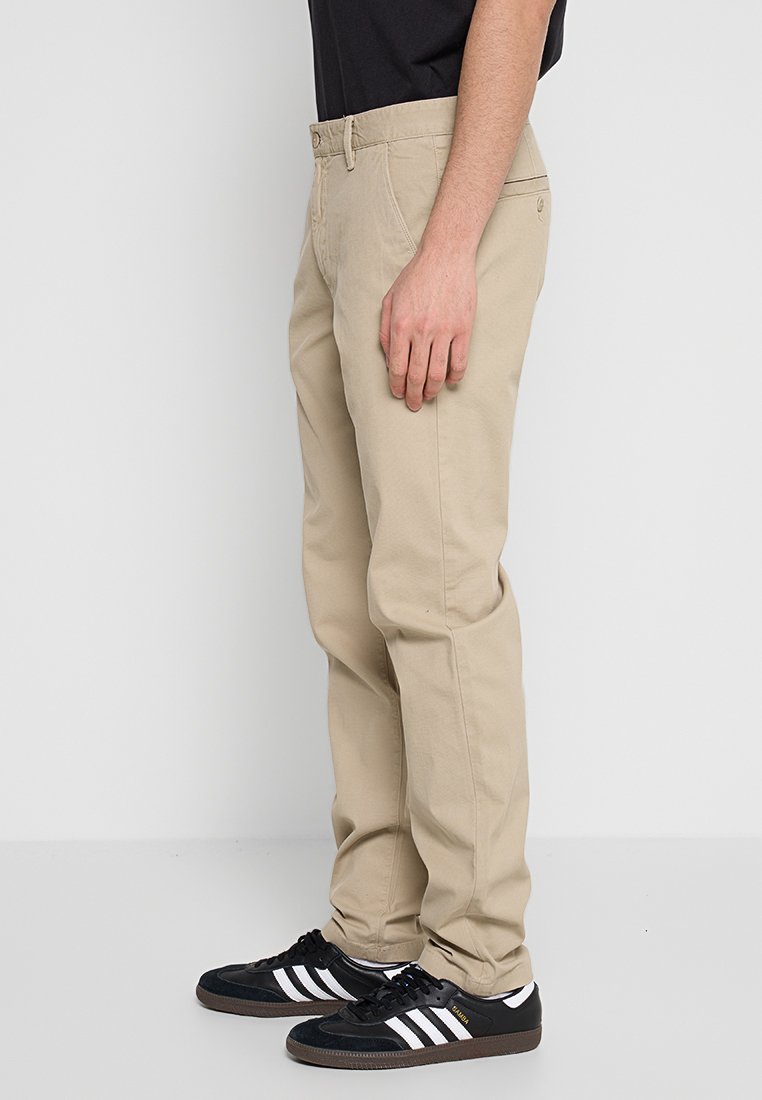 Beżowe bawełniane spodnie o prostym kroju, z bocznymi kieszeniami i szlufkami na pasek, w połączeniu z czarnymi sneakersami z białymi paskami.
