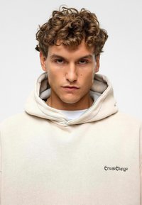 Beige katoenen hoodie met een voorzak, een trekkoordcapuchon en "CreateChange" geborduurd in het zwart op de linkerborst.