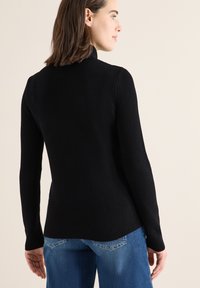 Maglione nero a collo alto a coste con maniche lunghe, design aderente, texture morbida e orlo leggermente curvo, abbinato a jeans blu.