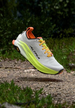 Chaussure de trail running gris clair et vert citron avec des accents orange, en l'air au-dessus d'un chemin de gravier entouré de feuillage vert.