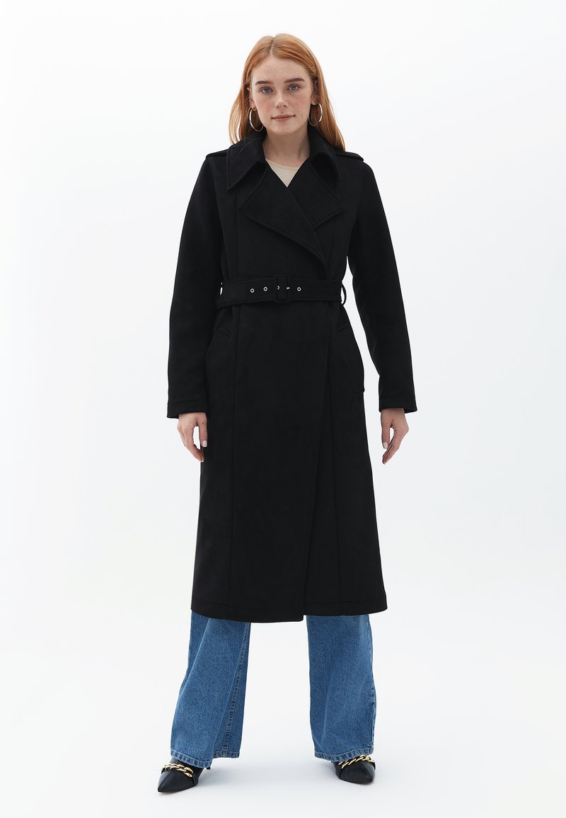 Cappotto trench nero con design doppiopetto, ampi revers, vita con cintura e tasche laterali, abbinato a jeans in denim a gamba larga.