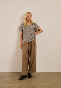 Femme blonde debout contre un mur beige, portant une blouse à manches courtes à motifs, un pantalon ample marron et des mocassins noirs, avec une main sur la hanche.