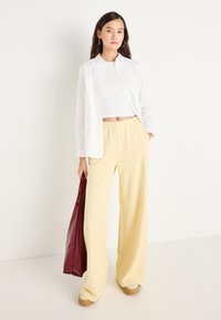 Ανοιχτό κίτρινο παντελόνι με φαρδιά πόδια και ελαστική μέση, συνδυασμένο με ένα λευκό πουκαμισάκι με κουμπιά και μια μπορντό δερμάτινη τσάντα.
