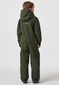 Grön vadderad overall med huva, med blixtlåsförsedda fickor, svarta detaljer på muddarna och en logga på ryggen. Lämplig för vinteraktiviteter.