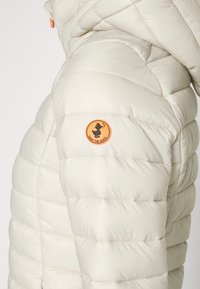 Cremefarvet pufferjakke med quiltet tekstur, der har et rundt orange badge mærket "SAVE THE DUCK" på den øverste ærme.