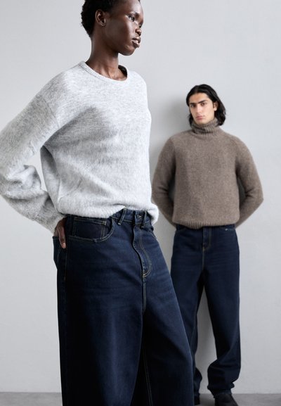 Gray textured sweater, dark denim wide-leg jeans with yellow stitching, featuring a high waist. A brown turtleneck sweater is worn in the background.