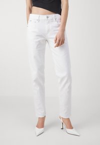 Bílé džíny z denimu s přímým střihem, pět kapes, zapínání na knoflíky a lehké opotřebení. V kombinaci s bílými botami na vysokém podpatku.