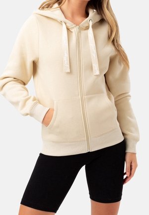 Femme portant un sweat à capuche beige zippé avec poches et des shorts de cycliste noirs sur un fond uni.