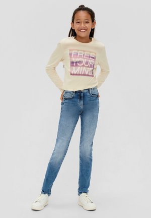 Crème lange mouwen shirt met paarse graphic "FREE YOUR MIND," gecombineerd met lichtblauwe jeans en witte sneakers, wat een casual look oplevert.