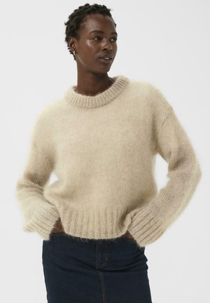 Beiger Strickpullover aus weichem, flauschigem Material, mit rundem Ausschnitt, langen Ärmeln und geripptem Saum, getragen über dunklen Jeans.