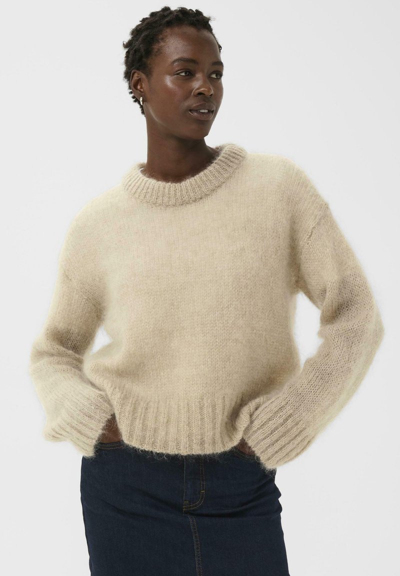 Pull beige en tricot fabriqué en matériau doux et duveteux, avec un col rond, des manches longues et un ourlet côtelé, porté sur un jean foncé.