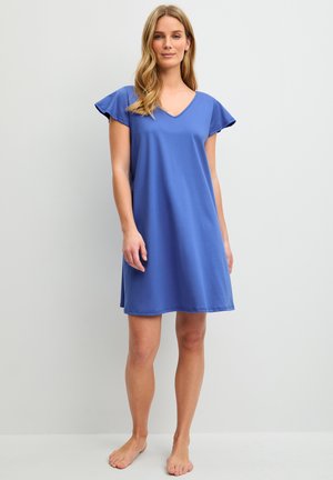 Femme debout pieds nus portant une robe bleue ample à manches courtes avec un col en V, sur un fond uni gris clair.