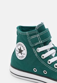Zielone wysokie trampki z płótna z białymi akcentami, okrągłym logo Chuck Taylor i metalowymi oczkami. Posiadają białe sznurowadła i teksturowaną tkaninę.