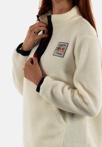 Maglione in fleece, colore crema, con collo zip, rifiniture blu navy. Presenta un logo quadrato con palme e dettagli solari sul lato sinistro del petto.