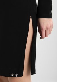 Svart klänning med hög slits, dragkedjedetalj på sidan och metalliska accenter vid fållen, tillverkad av mjukt, stretchigt material.