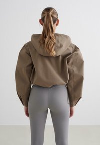 Brązowa kurtka z kapturem i oversize'owymi rękawami, zestawiona z szarymi legginsami. Kurtka ma gładką teksturę i minimalistyczny design.