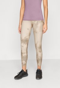 Ελαφριά, μπεζ κολάν με μοτίβο tie-dye και υφή, συνδυασμένα με μαύρα αθλητικά παπούτσια και εφαρμοστό μωβ τοπ.