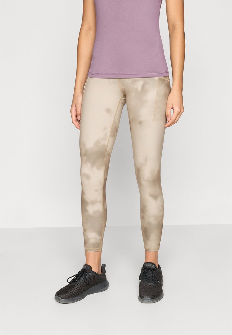 Ελαφριά, μπεζ κολάν με μοτίβο tie-dye και υφή, συνδυασμένα με μαύρα αθλητικά παπούτσια και εφαρμοστό μωβ τοπ.