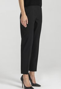 Kobieta ubrana w czarne, dopasowane spodnie do kostek i czarne, spiczaste szpilki, stojąca na tle gładkiego, białego tła.