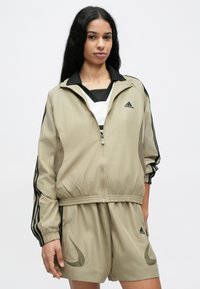 Bežová športová bunda s čiernymi akcentmi, predným zipsom a elastickým pásom, doplnená o zladené šortky s logom a zaoblenými detailmi na vreckách.