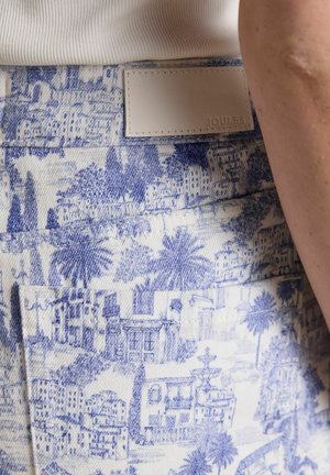 Close-up of blue and white patterned fabric with detailed buildings and palm trees, featuring a white leather label embossed with "Joules."