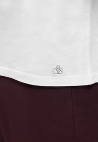 Άκρη λευκού πουκαμίσου με μικρή γκρι κεντημένη αγκύλη επάνω σε ύφασμα σκούρου μπουρντό παντελονιού.