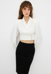 Blusa blanca corta con manga abullonada y detalle de tres botones, combinada con una falda lápiz negra ajustada. Textura de tela suave.