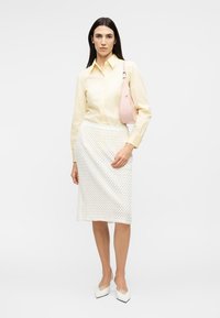 Hosszú sötét hajú nő világos sárga gombos ingben, texturált fehér térdharisnyában, fehér magassarkú cipőben, és egy rózsaszín válltáskát visel.