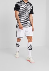 Camiseta de fútbol a rayas blancas y negras con mangas cortas, combinada con pantalones cortos blancos y calcetines con acentos negros. Presenta el logo de adidas y bordados.
