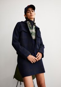 Marineblauwe wollen jas met twee vo pockets, gelaagd over een groen geruit shirt, afgewerkt met een zwarte coltrui en een marineblauwe pet.