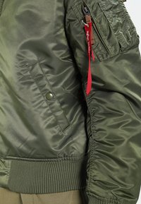 Olive green bomber jacket with a smooth, shiny finish, featuring ribbed cuffs and a zippered pocket with a red pull tab.