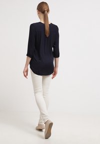 Mörkblå blus med långa ärmar i ett mjukt tyg och rundad nederkant, i kombination med vita slim fit-byxor och ljusa ballerinatöjor.