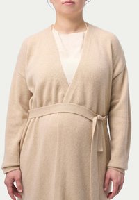 Ljusbeige kardiagan gjort av mjukt material, med djup V-ringning, långa ärmar och ett knytbälte i midjan. Slät textur, enkel design.