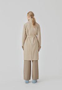 Beige trench coat with a belted waist, long sleeves, and a collar, paired with loose beige pants and blue heeled shoes. Back view shown.