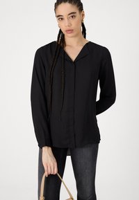 Blouse noire à manches longues avec un col à revers, texture lisse, poignets boutonnés et fentes latérales. Porte avec un jean foncé et tient un sac à main beige.