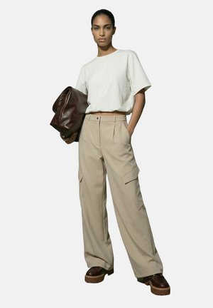 Kvinne iført hvit kort T-skjorte, beige vide cargobukser, brune kraftige sko, holder en stor brun skinnclutch.