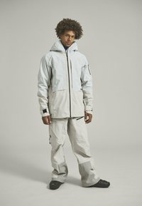 Light gray waterproof jacket with a zip front, large pockets, and adjustable hood, paired with matching cargo pants featuring paneled design and zippers.
