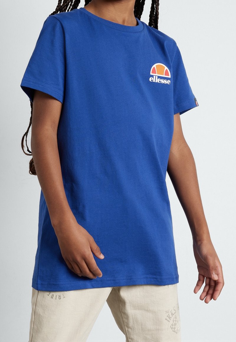 Enfant portant un t-shirt Ellesse bleu avec un petit logo sur la poitrine et un pantalon beige clair, debout devant un fond uni.