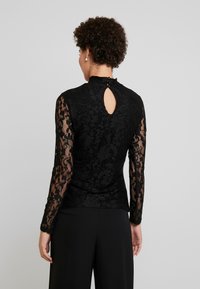 Svart spetsblus med långa ärmar, hög krage och nyckelhål bak med knappdetalj. Designad med blommönster och strukturerad tyg.