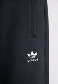 Black cotton sweatpants featuring a white embroidered Adidas logo with three stripes above the leaves, smooth texture, and tapered design.