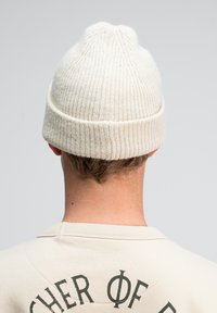 Cremfarbener Strickbeanie mit gerippter Textur und einem leichten Punkt an der Spitze, getragen mit einem beigefarbenen Sweatshirt mit bedrucktem Text auf dem Rücken.