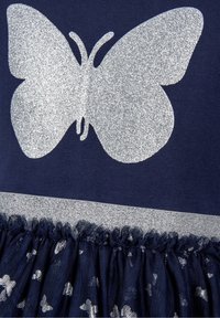 Námořnické šaty s velkým stříbrným třpytivým motivem motýla, s lesklým stříbrným pasem a vrstvenou tylovou sukní zdobenou malými stříbrnými hvězdami.