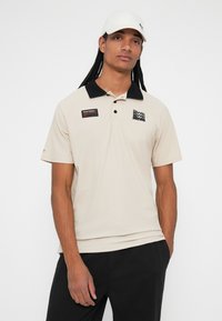 Beige polo shirt with a black collar, featuring a logo patch on the chest, short sleeves, and a smooth fabric texture.