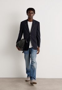 Blazer preto com costura branca, vestido sobre uma camisa branca, combinado com jeans azuis desbotados e ténis cinzentos. Segurando uma mala de mensageiro preta.