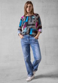 Svart topp med geometriska mönster i blått, rosa och vitt; matchad med ljusblå jeans och vita sneakers. Avslappnad passform och uppvikta ärmar.